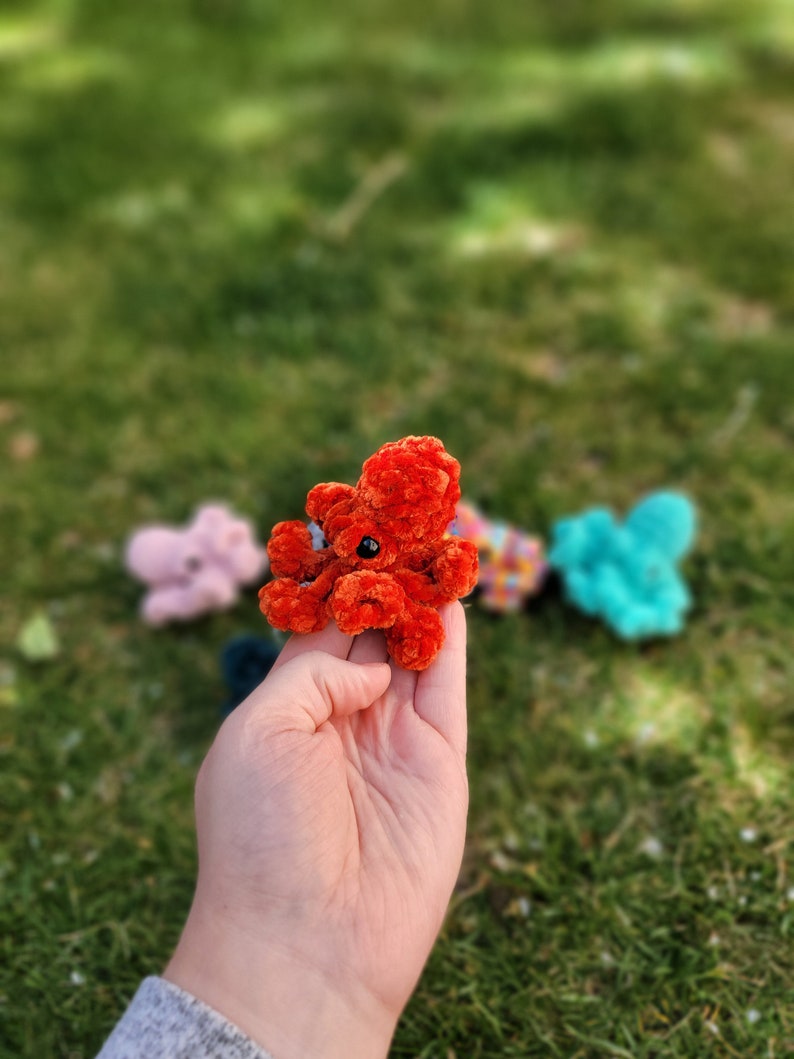 Puede incluir: Un peque&ntilde;o pulpo de crochet hecho de hilo rojo con ojos negros. El pulpo est&aacute; siendo sostenido en la mano de una persona.