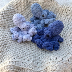 Puede incluir: Tres peluches de pulpo tejidos a crochet en tonos de azul, azul claro y morado, descansando sobre una manta de crochet beige. La manta tiene un borde con flecos. El texto "@bluejayehhandmade" es visible en la esquina superior derecha de la imagen.