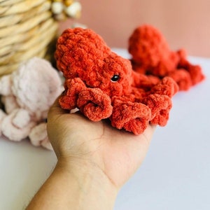 Puede incluir: Un juguete de peluche de pulpo naranja tejido a crochet con ojos negros. El pulpo está sentado en la mano de una persona.