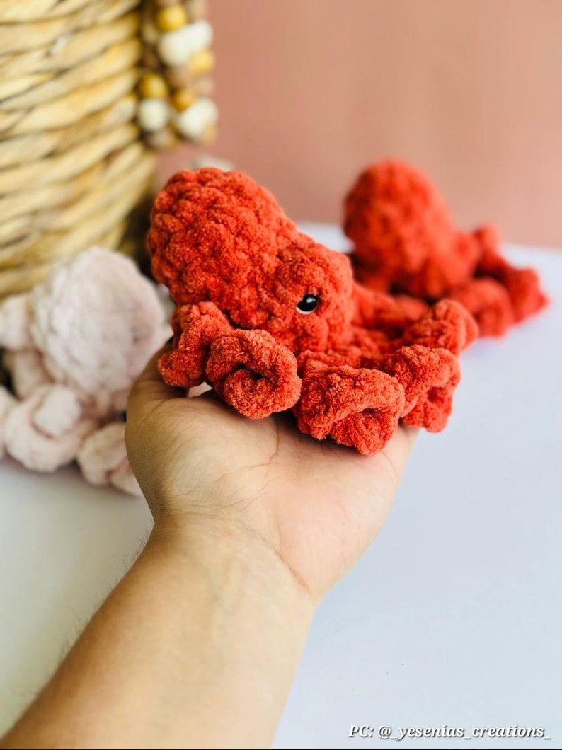 Puede incluir: Un juguete de peluche de pulpo naranja tejido a crochet con ojos negros. El pulpo est&aacute; sentado en la mano de una persona.