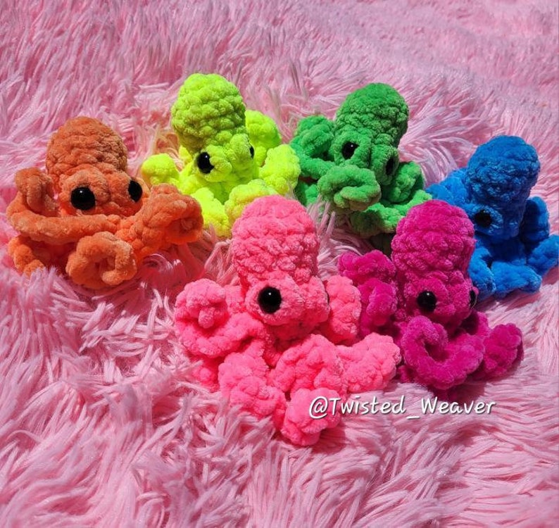 Puede incluir: Seis peluches de pulpo tejidos a crochet en varios colores brillantes, incluyendo naranja, verde, rosa y azul. Los peluches est&aacute;n dispuestos sobre una superficie esponjosa rosa. El texto "@Twisted_Weaver" es visible en la esquina inferior derecha de la imagen.