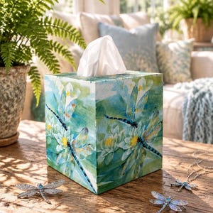 May include: A square tissue box cover featuring a painted scene of blue dragonflies and white water lilies on a green and blue background. Two silver-toned dragonfly brooches with blue accents are placed on the wooden surface nearby.