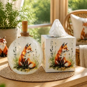 May include: A round glass bottle with a wooden stopper and a square tissue box, both featuring a printed illustration of a fox sitting in a forest setting with birch trees and green foliage. The items are placed on a round woven placemat.