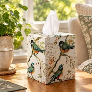 May include: A square tissue box cover featuring a cream-colored background with a printed pattern of small birds perched on brown branches with green leaves and berries, accented by faint, cursive script text across the surface.