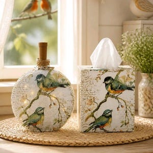 May include: A round glass bottle with a cork stopper and a rectangular tissue box, both featuring a white background with a printed design of blue and orange birds perched on branches, accented with faint script text and floral patterns.