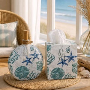 May include: A decorative set featuring a round vase with a wooden stopper and a tissue box. Both items are white with a sea-themed design of blue starfish, seashells, and coral. The set is placed on a woven mat.