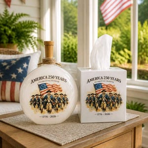 May include: A round white glass bottle with a wooden stopper and a square white tissue box, both featuring a graphic of soldiers carrying an American flag with the text AMERICA 250 YEARS CELEBRATING FREEDOM 1776-2026 printed in black ink.