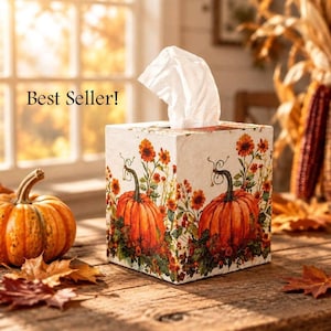 May include: A square tissue box cover featuring a white background with a printed design of orange pumpkins, green vines, and orange and yellow flowers. A white tissue is pulled through the top opening. The text Best Seller! is visible.