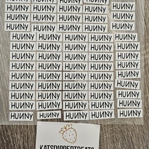 May include: A collection of small, rectangular white labels with the word "HUNNY" printed in black, arranged on a clear sheet. A separate label features a strawberry graphic and the text "KATSDIPPEDTREATS Katherine Hernandez."