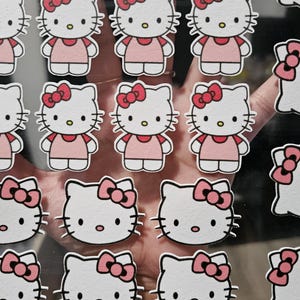 May include: A collection of Hello Kitty stickers. The stickers feature the iconic character with a white face, a red bow, and a pink dress. The stickers are arranged on a clear sheet, ready for use.