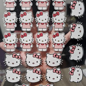 May include: A collection of Hello Kitty stickers. The stickers feature the iconic cartoon cat in a pink dress and bow. Some stickers are just the cat's face with a bow. The stickers are white, pink, and black.