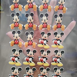 May include: A collection of Mickey Mouse stickers. The stickers feature Mickey in various poses, wearing his signature red shorts, yellow shoes, and white gloves. The stickers are on a clear sheet.