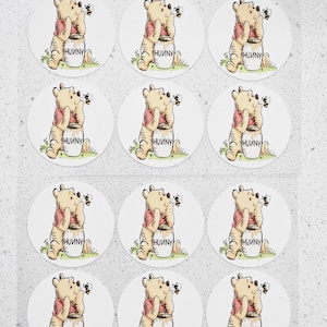 May include: A sheet of round stickers featuring a cartoon bear in a red shirt, sitting next to a honey pot. The honey pot has the word "HUNNY" printed on it. A small bee is flying near the bear.