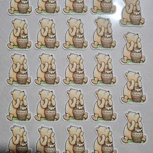 May include: A sheet of twenty-four die-cut stickers featuring a light yellow bear sitting on green grass next to a brown honey pot labeled HUNNY. The bear is depicted in a seated position with one hand resting on its chin, looking upward.