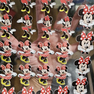 May include: A collection of Minnie Mouse stickers and decals. The stickers feature Minnie Mouse in her classic red and white polka dot bow, skirt, and yellow shoes. Some stickers show Minnie in a standing pose, while others show her face.