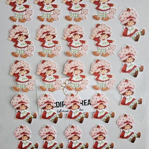 May include: A collection of Strawberry Shortcake character stickers. The stickers feature the character in a red dress, a strawberry hat, and green and white striped leggings. Some stickers include a small pink bear.