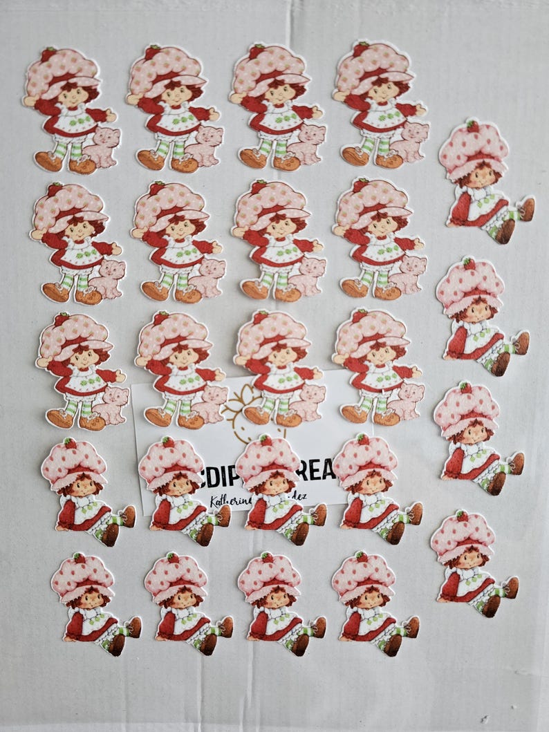 May include: A collection of Strawberry Shortcake character stickers. The stickers feature the character in a red dress, a strawberry hat, and green and white striped leggings. Some stickers include a small pink bear.
