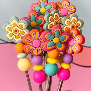 May include: A set of six colorful flower-shaped drink stirrers with colorful beads. The stirrers are made of metal and have a flower-shaped top with a colorful design. The beads are made of plastic and are in a variety of colors, including pink, yellow, orange, and purple.
