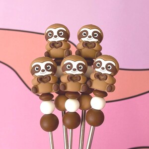 May include: Six decorative skewers featuring brown sloth figurines holding heart shapes. Each skewer has a series of brown and white beads, with a stainless steel stick. The background is a pink and purple color.
