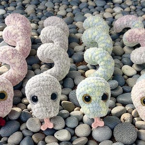 May include: Four handmade crochet snake toys in various pastel colors, including pink, gray, and blue. Each snake has large, round eyes and a small, protruding tongue. The snakes are arranged on a bed of smooth, gray and blue stones.