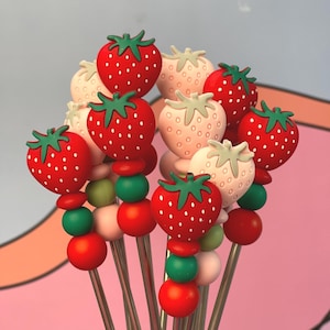 May include: A set of eight stainless steel stirring straws with red and green silicone beads and strawberry-shaped toppers. The strawberries are red and white with green leaves.