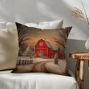 May include: A decorative throw pillow featuring a winter scene with a red barn, snow-covered ground, and bare trees. The pillow has a neutral background and is placed on a white sofa. A second pillow with a tan cover is also visible.