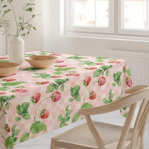 May include: A pink and white checkered tablecloth with a repeating pattern of strawberries and green leaves. Two wooden bowls, a white pitcher, and a vase with greenery sit on the table. A wooden chair is in the foreground.