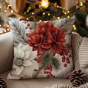 May include: Square decorative pillow with a floral design featuring a large red flower and a white flower, with green pine needles and red berries. The pillow is on a beige couch with a cozy blanket and pine cones.