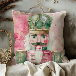 May include: A decorative square pillow featuring a watercolor illustration of a nutcracker soldier. The pillow has a pink background with a splash design. The nutcracker is in shades of pink, green, and gold, with a white beard and mustache.