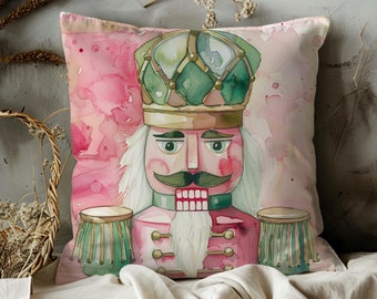 May include: A decorative square pillow featuring a watercolor illustration of a nutcracker soldier. The pillow has a pink background with a splash design. The nutcracker is in shades of pink, green, and gold, with a white beard and mustache.