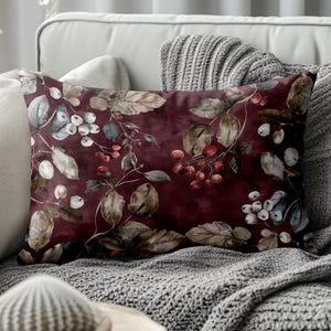 May include: A rectangular decorative pillow with a burgundy background and a botanical print. The print features brown leaves, white berries, and red berries. The pillow is placed on a gray sofa with a gray knitted blanket.