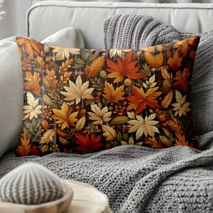 May include: Rectangular decorative pillow featuring a fall leaves pattern. The pillow displays a variety of autumn leaves in shades of red, orange, yellow, and brown against a dark background. The pillow is placed on a gray sofa with a gray blanket.