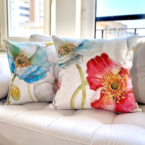 May include: Two decorative throw pillows with floral patterns. One pillow features a blue poppy with green stems and leaves on a white background. The other pillow features a red poppy with yellow center and green stems and leaves on a white background.