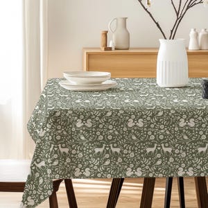May include: A green tablecloth with a white woodland pattern featuring deer, birds, and floral elements. The tablecloth is draped over a wooden table, with a wooden chair visible on the left. A white vase holds branches with white flowers.