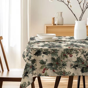 May include: A rectangular tablecloth with a holly and berry pattern in green, red, and cream covers a wooden table. A wooden chair is on the left. A white vase holds branches with white flowers. White plates and bowls are on the table.