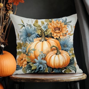 May include: A decorative throw pillow featuring a watercolor design of pumpkins and flowers in shades of orange, blue, and yellow. The pillow is placed on a wooden stool, with pumpkins in the foreground.