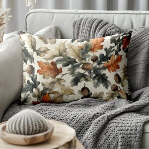 May include: Rectangular decorative pillow featuring a watercolor design of oak leaves and acorns in shades of orange, brown, and green on a cream background. The pillow is placed on a gray sofa with a matching throw blanket, creating a cozy, autumnal theme.