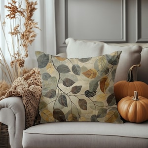 May include: Square throw pillow with a botanical leaf pattern in shades of gray, brown, and yellow. The pillow is on a beige sofa, with two orange pumpkins nearby. The pillow's design features a network of branches and leaves.