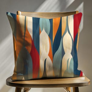 May include: Square decorative pillow with an abstract design. The pillow features a pattern of vertical shapes in shades of red, orange, blue, and cream. The pillow is placed on a wooden chair.