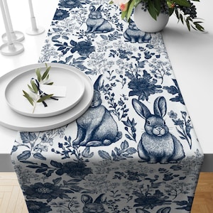 May include: A white table runner with a blue floral and bunny rabbit pattern. The runner features a repeating pattern of blue flowers, leaves, and bunnies.