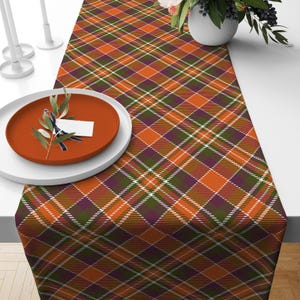 May include: Orange, green, and purple plaid table runner with a white background. The runner is on a table with a white plate and a sprig of greenery.