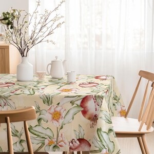 May include: A floral tablecloth with a cream background and a pattern of large pink and red flowers with green leaves. A white teapot, vase, and cups are on the table. Wooden chairs surround the table.