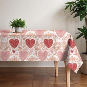 May include: A white tablecloth with a pink and red heart pattern. The tablecloth is laid on a wooden table with a potted plant on top.
