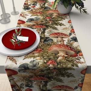 May include: A white table runner with a repeating pattern of red and white mushrooms, green ferns, and brown leaves. The runner is on a table with a red plate and a sprig of greenery.