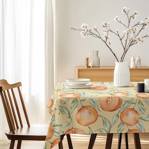 May include: A rectangular tablecloth with a repeating pattern of oranges and green leaves on a light yellow background. The table is set with white plates and a wooden chair is visible on the left. A white vase holds flowering branches.