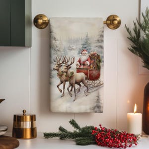 May include: A white kitchen towel with a watercolor illustration of Santa Claus riding in a red sleigh pulled by two reindeer through a snowy forest. The reindeer are adorned with green garlands.