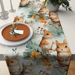 May include: A white table runner with a repeating pattern of brown squirrels, green leaves, and orange and yellow fall leaves.
