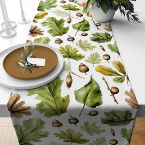 May include: A white table runner with a repeating pattern of green and brown leaves and acorns. The leaves are in various shades of green and brown, and the acorns are a light brown color.