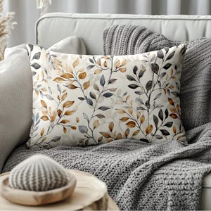 May include: Rectangular decorative pillow with a botanical design. The pillow features a cream-colored background with a repeating pattern of stylized branches and leaves in shades of brown, gray, and blue. The pillow is placed on a gray sofa with a matching throw blanket.