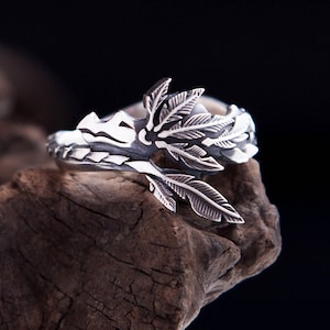May include: A silver ring featuring a detailed design of overlapping leaves and branches that wrap around the band. The metal has a polished finish with darkened recessed areas to highlight the intricate texture of the foliage motifs.